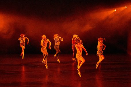 Tanzende Personen in leuchtenden Kostümen auf einer Bühne mit orangefarbenem Licht.