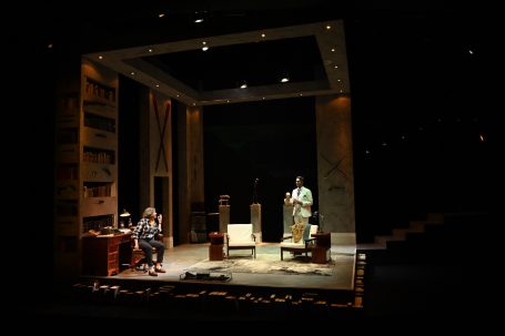 Bühne mit zwei Schauspielern in einem Wohnzimmer-Szenario, Bücherregale im Hintergrund.