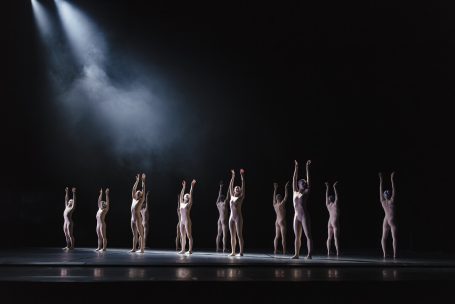 Tanzensemble in eleganten Outfits bewegt sich unter einem Lichtstrahl auf der Bühne.