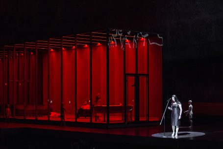 Bühnenbild mit rotem Licht, zwei Figuren in einem transparenten Raum.
