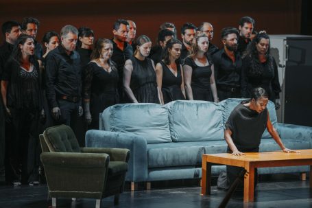 Eine Sängerin sitzt am Tisch, während ein Chor im Hintergrund auftritt.