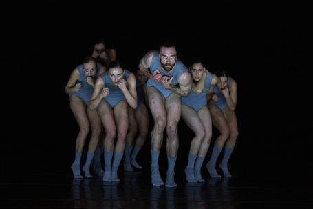 Gruppentanz mit mehreren Tänzern in blauer Kleidung vor dunktem Hintergrund.