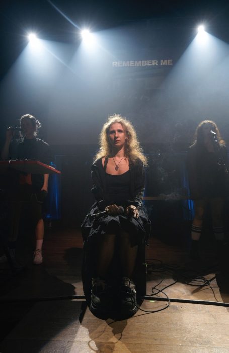 Frau sitzt im Mittelpunkt auf der Bühne, umgeben von Schatten und schwachem Licht.