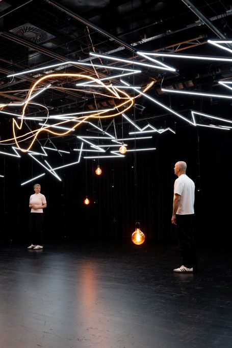 Zwei Personen in einem dunklen Raum mit leuchtenden Linien und Hängelampen.