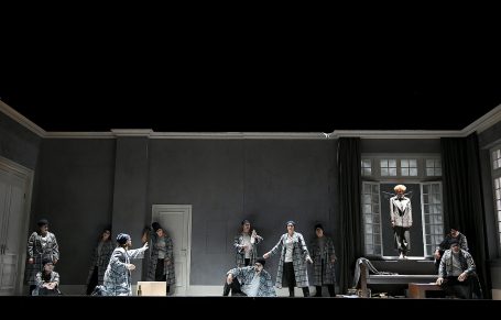 Eine Theaterbühne mit mehreren Personen in grauen Kostümen, die eine Inszenierung aufführen.