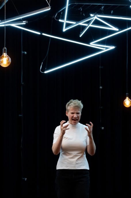 Eine Person in einem weißen Oberteil steht vor einer dunklen Wand mit geometrischen Lichtern.