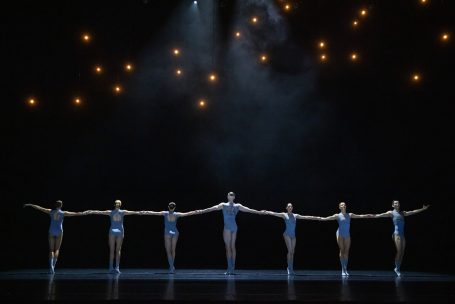 Sechs Balletttänzer in blauen Kostümen stehen in einer Linie auf der Bühne.
