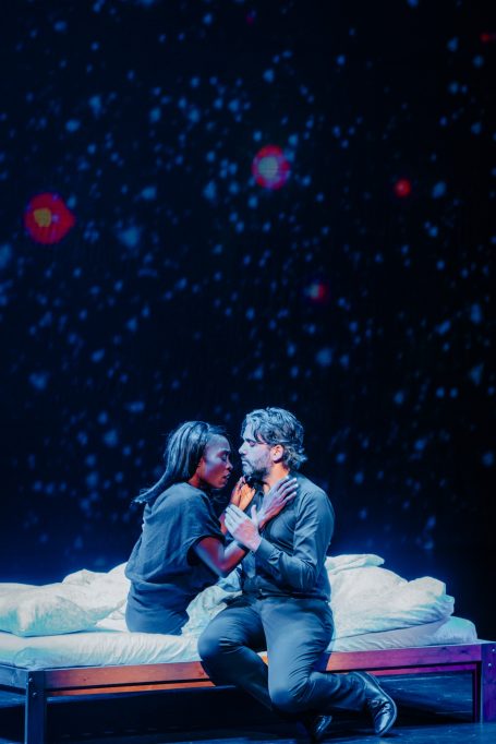 Zwei Personen sitzen auf einem Bett und schauen sich verliebt an, vor einem nächtlichen Himmel.