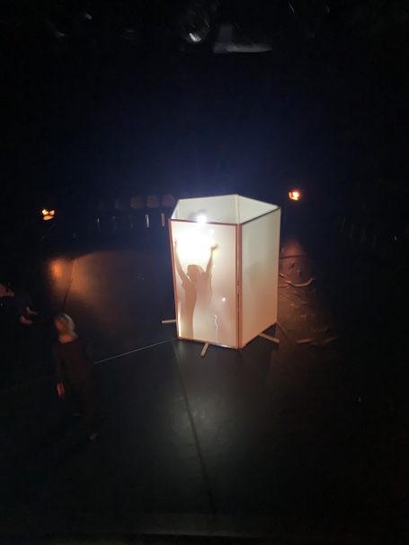 Eine beleuchtete Box auf einer dunklen Bühne, mit einer Silhouette im Inneren.