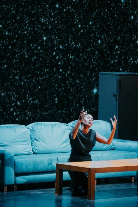 Eine Tänzerin in schwarzem Kleid, kniend vor einem Tisch, vor einem blauen Sofa.