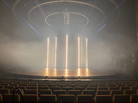 Bühne mit Lichtern in einem nebligen Theater, leere Sitzreihen im Vordergrund.