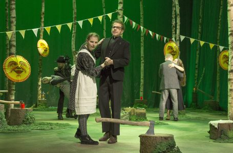 Zwei Personen tanzen im Wald, umgeben von Festdekoration und weiteren Personen im Hintergrund.