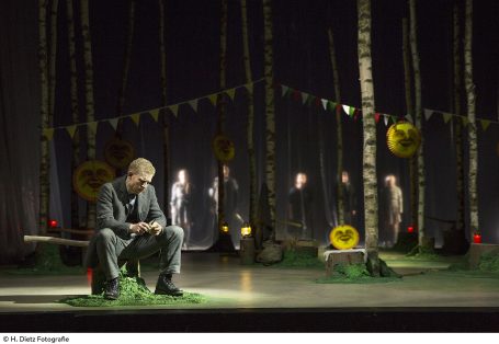 Ein Mann sitzt nachdenklich im Wald, während Figuren im Hintergrund sichtbar sind.