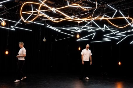 Zwei Tänzer in einem beleuchteten Raum mit geometrischen Lichtinstallationen.