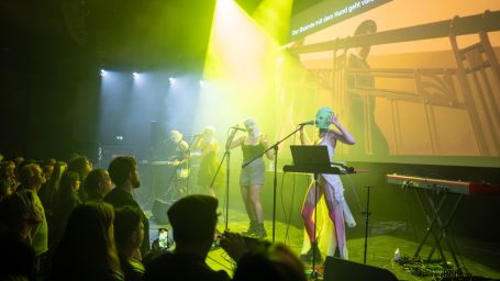 Bunt beleuchtete Musikperformance mit Darstellern auf der Bühne vor einem Publikum.