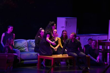 Gruppe von Personen auf einer Bühne, gekleidet in Schwarz, versammelt um einen Holztisch.